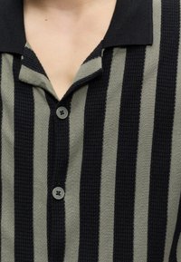 Κοντινό πλάνο ενός ατόμου που φοράει μια μαύρη και γκρι κάθετα ριγέ πουκάμισο με γιακά και δύο ορατά κουμπιά.