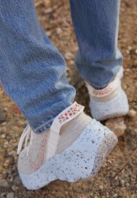 Světle šedé sportovní boty s bělavou podrážkou posetou tečkami, vícebarevným vzorem po stranách a texturovaným svrškem. Nošené s modrými džínami.