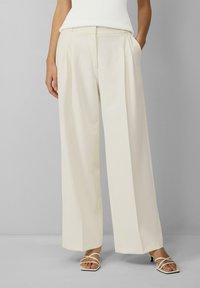 Crème wijde broek met een gladde textuur, geplooide voorkant en zijzakken, gecombineerd met witte sandaaltjes met bandjes.