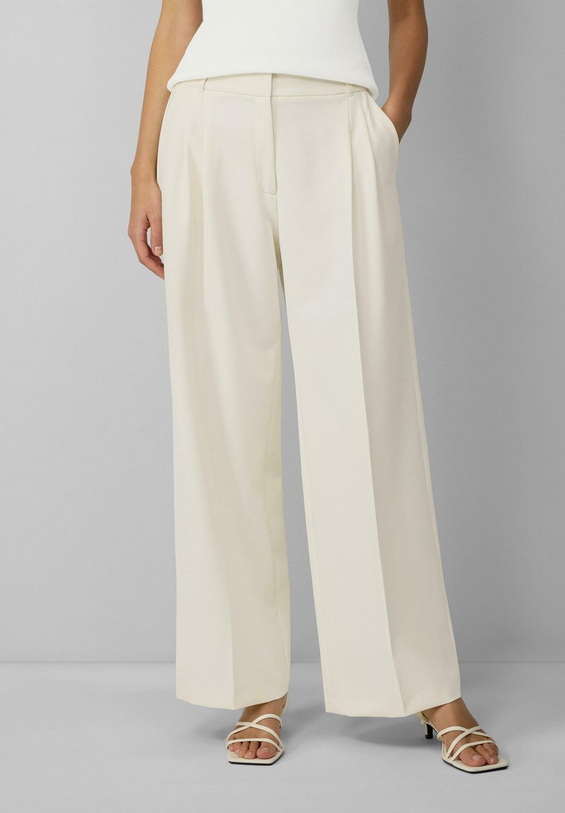 Crème wijde broek met een gladde textuur, geplooide voorkant en zijzakken, gecombineerd met witte sandaaltjes met bandjes.