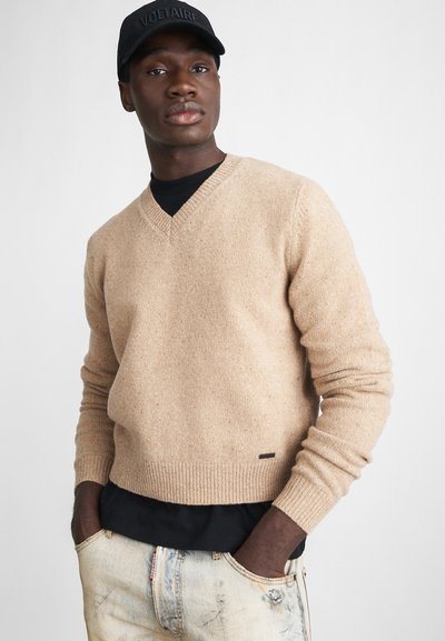 Maglione lavorato a maglia beige con scollatura a V, polsini e orlo a costine. Indossato sopra una camicia nera e abbinato a jeans azzurri chiari strappati.