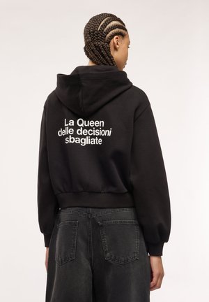 Donna con capelli intrecciati che indossa una felpa nera con cappuccio con testo italiano "La Queen delle decisioni sbagliate" e jeans neri, vista da dietro.