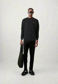 Sweater preto com decote redondo, logo bordado no peito, combinado com calças pretas e uma mala tote preta acolchoada. Ténis pretos.