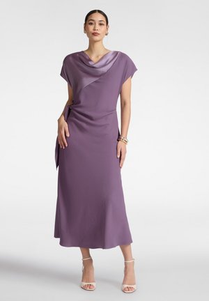 Frau steht in einem ärmellosen, knöchellangen lavendelfarbenen Kleid mit drapiertem Ausschnitt und asymmetrischem Detail, trägt weiße High Heels und Armbänder.