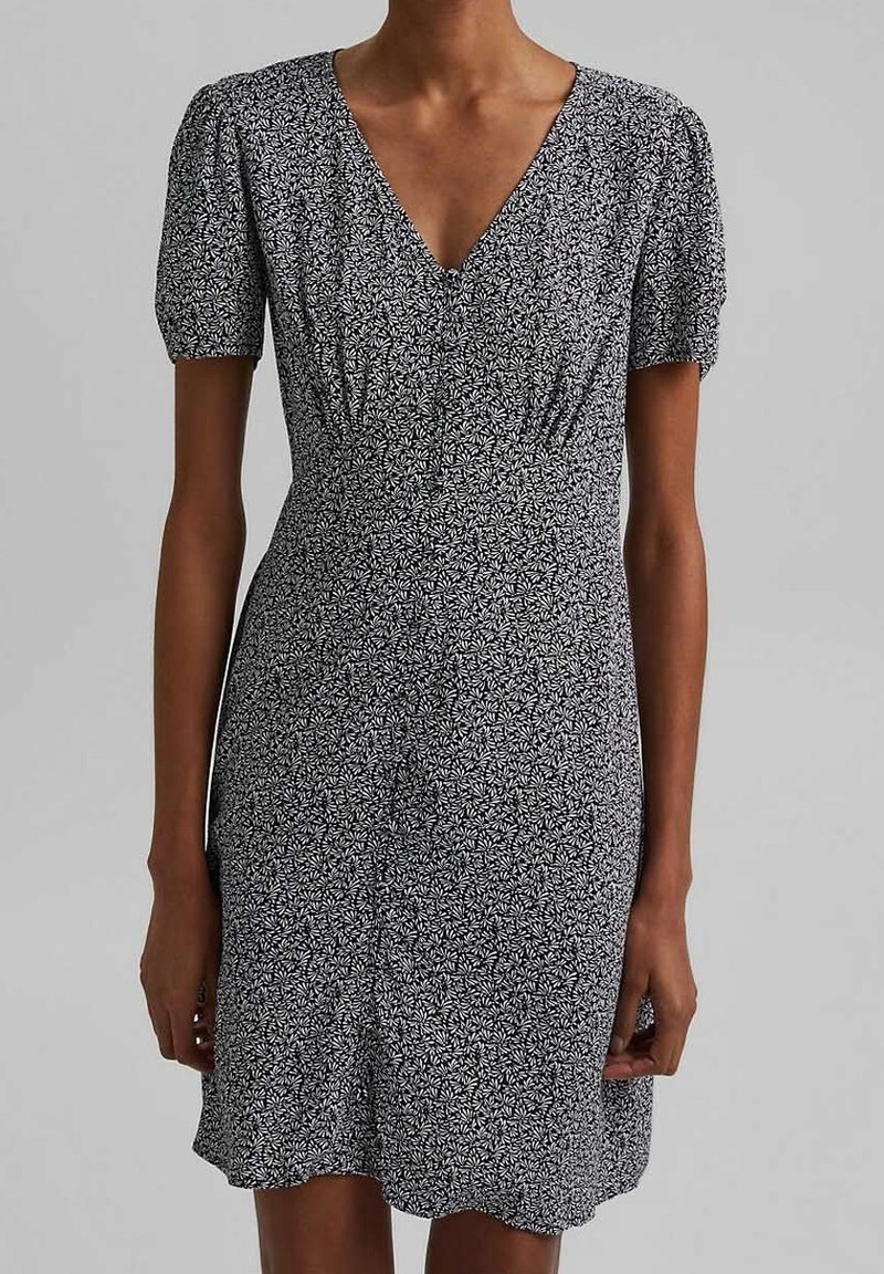Femme portant une robe noire à manches courtes avec un motif floral blanc et un décolleté en V, debout devant un fond uni gris clair.