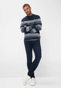 Maglione a righe blu navy e grigio realizzato in tessuto a maglia, abbinato a jeans di denim scuro e sneakers bianche. Il modello è in piedi con le braccia incrociate.