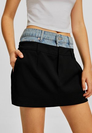 Femme portant une jupe noire superposée sur un jean en denim bleu visible à la taille, avec une main dans la poche de la jupe et un top court blanc.