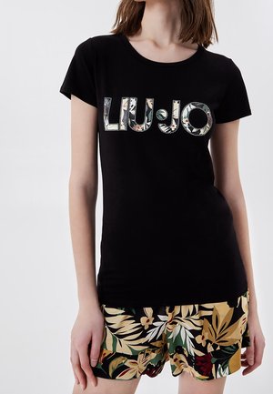 Kobieta ubrana w czarną koszulkę z krótkim rękawem z kwiatowym logo "Liu Jo" oraz krótkie spodenki w tropikalny wzór w kolorach zielonym, beżowym, czerwonym i czarnym.