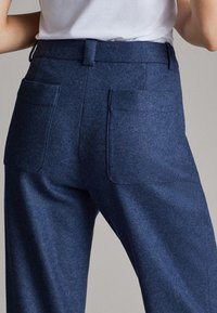 Vue de dos d'une personne portant un pantalon bleu droit avec de grandes poches arrière et une chemise blanche rentrée.