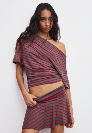 Jonge vrouw die een grijs-rood gestreepte off-shoulder top en bijpassende rok draagt, en met één hand de tailleband lichtjes optrekt.