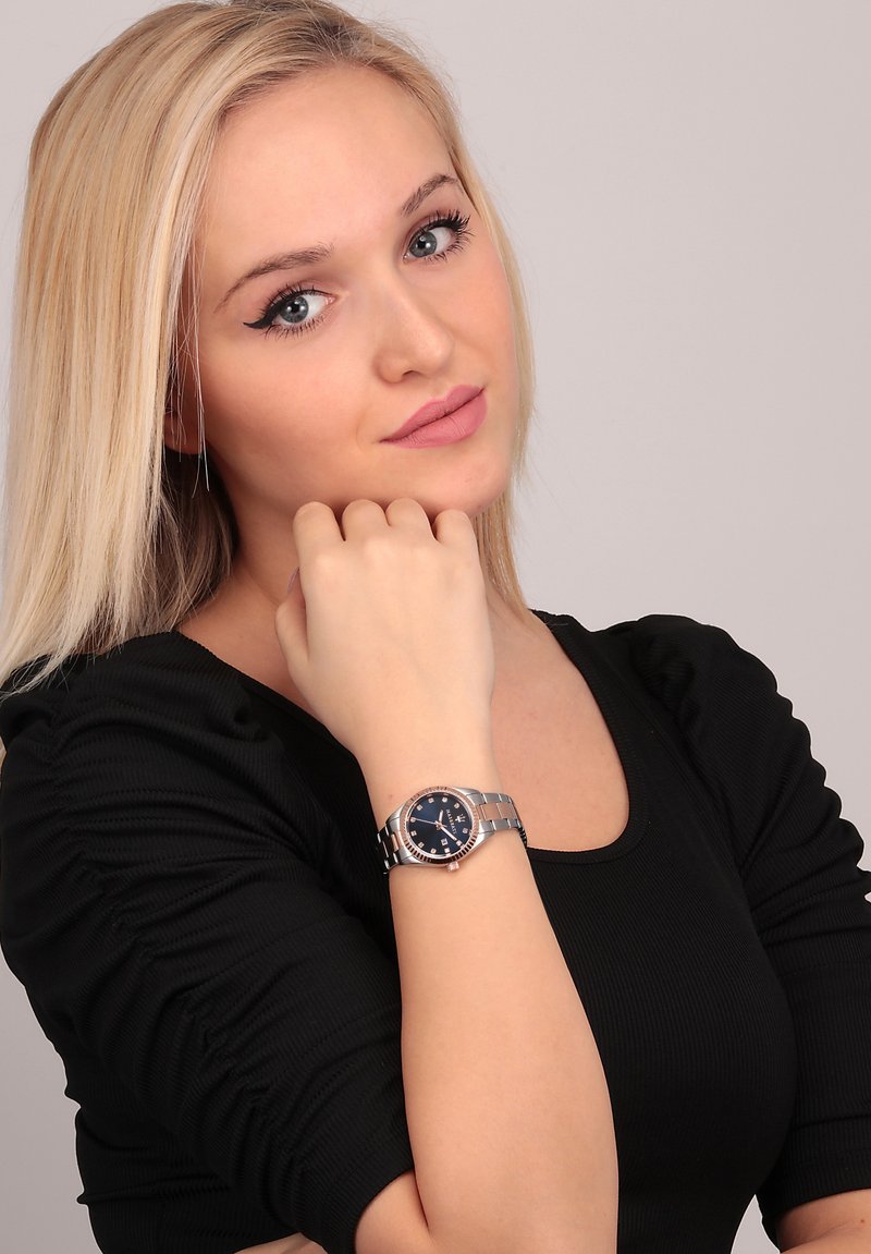 Orologio in acciaio inossidabile argentato con quadrante blu, con cassa rotonda e bracciale a maglie, indossato su un polso con una maglietta nera a trama.