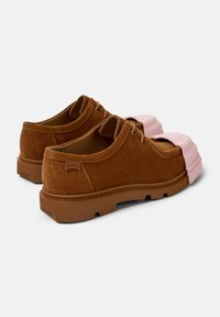 Scarpe stringate in suede marrone con suole spesse, dotate di distintive punte in gomma rosa, mostrate da una vista posteriore a tre quarti.