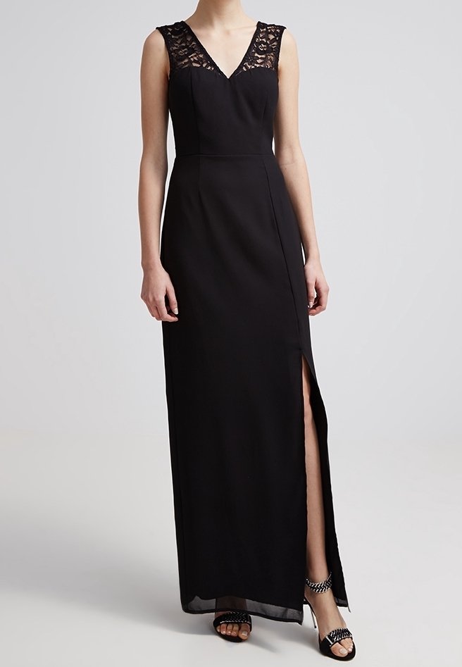 Robe de soirée noire sans manches avec décolleté en V en dentelle et fente latérale, portée avec des talons ouverts noirs cloutés.
