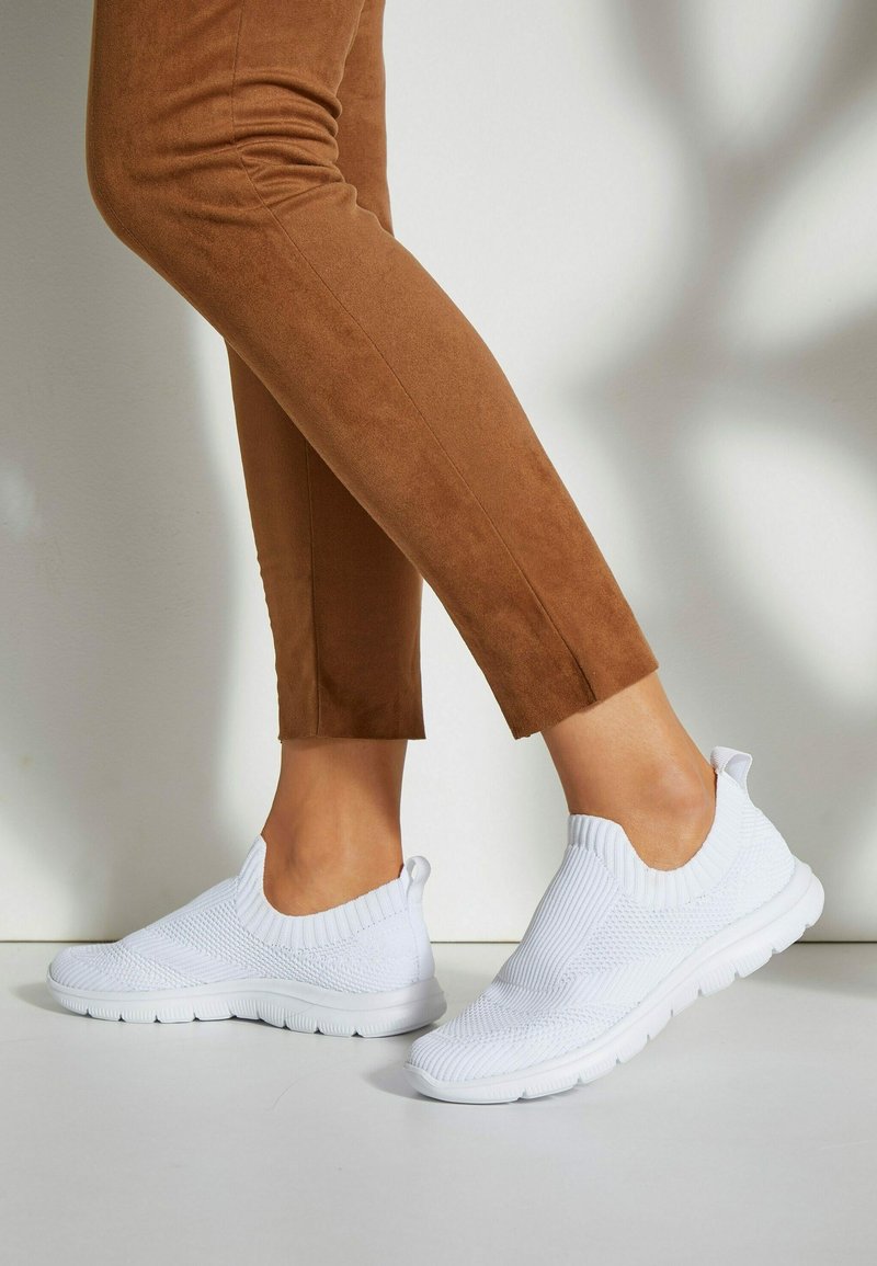 Weiße gestrickte Slip-On-Sneaker mit strukturierter Oberfläche und geripptem Kragen, kombiniert mit braunen Wildleder-Hosen, die einen lässigen Look zeigen.