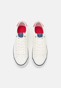 Zapatillas blancas con parte superior de cuero liso, detalles en azul marino y lengüetas en rojo en el talón. Presentan cordones blancos y suela en contraste azul marino.