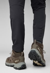 Scarpe da trekking marroni con suole grigie, caratterizzate da una combinazione di materiali scamosciati e in rete. Pantaloni neri imbottiti con una vestibilità aderente attorno alle caviglie.