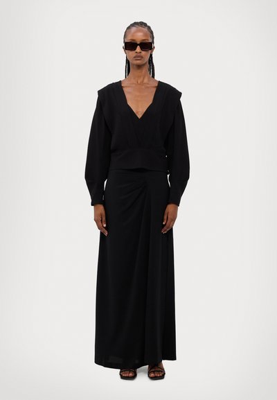 Black dress featuring a deep V-neck, long puff sleeves, and a cinched waist, with a flowing skirt and asymmetrical draping. Worn with dark sunglasses.