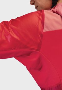 Rote Sportjacke mit einer Mischung aus glänzenden und matten Texturen. Hervorstechende Designelemente sind farblich abgesetzte Bereiche und gestreifte Akzente.