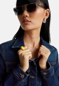 Džínová bunda s oranžovým prošíváním, průhledné sluneční brýle a žlutý prsten. Hladká textura, tmavě modrá barva, ukazuje moderní, přiléhavý design.