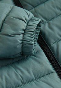 Padded jacket in muted teal, featuring a smooth texture, ribbed cuffs, and a black zipper detail along the front.