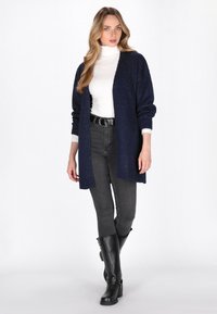 Marinblå stickad kofta med öppen framsida, ribbad mudd, bärs över en vit polotröja och grå slim-fit jeans, kombinerad med svarta knähöga stövlar.
