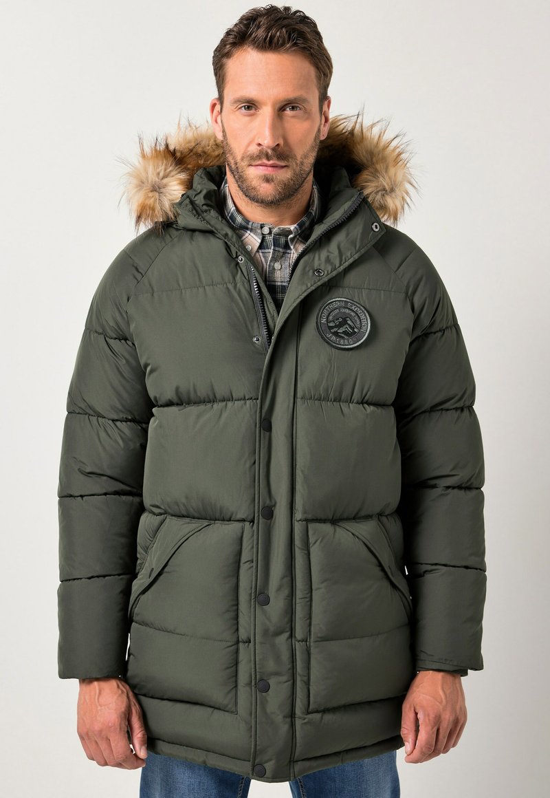 Homme portant une veste d'hiver matelassée verte avec capuche en fourrure et patch de logo de montagne, associée à une chemise à carreaux et un jean bleu.