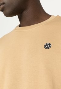Sudadera beige con cuello redondo, hecha de algodón suave, que presenta un pequeño logo circular negro en el pecho izquierdo. Textura lisa y ajuste casual.