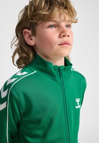 Băiat cu păr şaten deschis şi pistrui, purtând o geacă sport verde cu fermoar și dungi albe sub formă de chevron pe mâneci, privind ușor în sus.