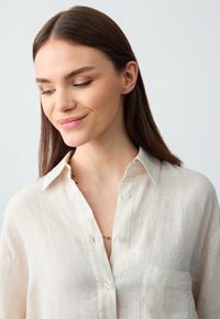 Camicia di lino beige con colletto, bottoni frontali e taschino sul petto. Texture liscia e materiale leggero, adatta per un look casual.