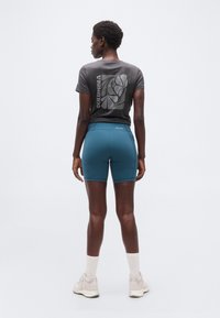 Γυναίκα με γκρι μπλουζάκι με σχέδιο, βερμούδα αθλητική τυρκουάζ, λευκές κάλτσες και μπεζ αθλητικά παπούτσια, που στέκεται με την πλάτη προς το μέρος μας σε απλό λευκό φόντο.