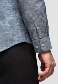 Main d'homme à côté du torse portant une chemise à manches longues grise à motif floral, avec des poignets à deux boutons, et un pantalon noir, sur un fond uni.