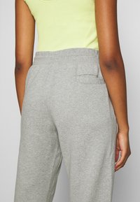 Grijze sweatpants met een ontspannen pasvorm, gemaakt van zachte stof. Heeft een elastische tailleband en een enkele achterzak. Heldere groene mouwloze top zichtbaar.