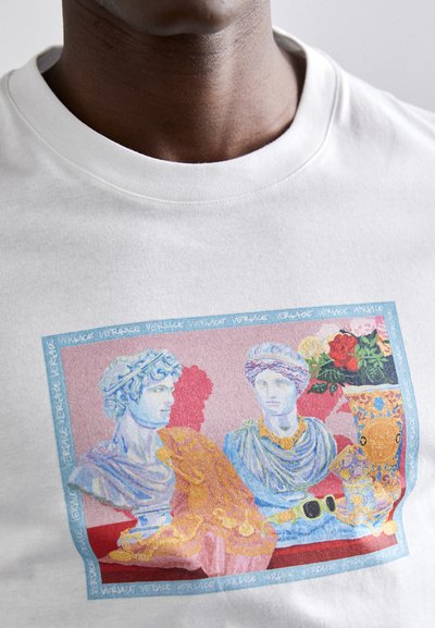 Acercamiento a una camisa blanca con un gráfico colorido de dos bustos clásicos, una cabeza de caballo decorativa, un jarrón con flores y unas gafas de sol sobre un fondo rojo.