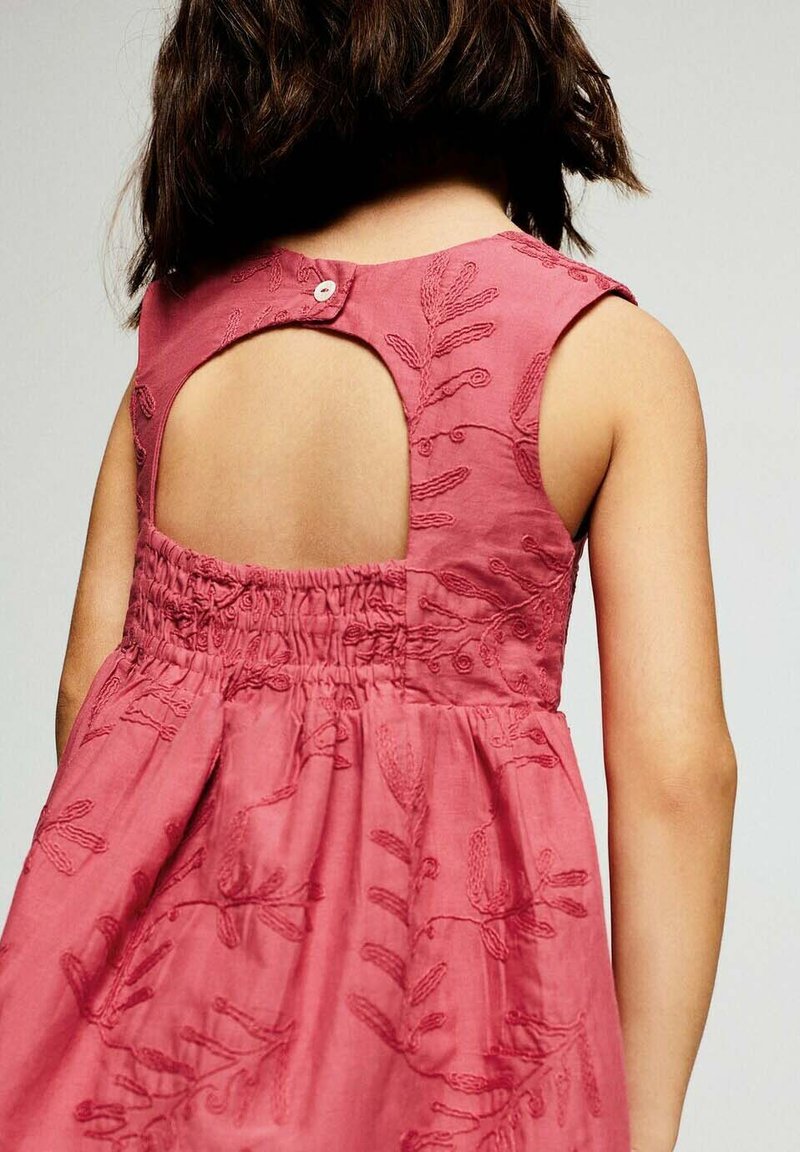 Enfant aux cheveux bruns mi-longs portant une robe rose sans manches avec des motifs de feuilles brodés et un dos avec une ouverture en forme de goutte maintenue par un bouton.