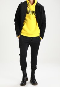 Černá bunda na zip s texturovanou látkou přes jasně žlutou mikinu s černým nápisem. Černé zúžené kalhoty a černé šněrovací boty.