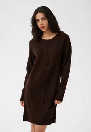 Femme aux cheveux foncés et ondulés, portant une longue robe pull en tricot marron avec des poignets côtelés et une couture centrale, debout devant un fond uni.