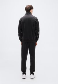 Μαύρη φόρμα που περιλαμβάνει ζακέτα με φερμουάρ και ψηλό γιακά με άσπρη ρίγα, συνδυασμένη με παντελόνι στο ίδιο σχέδιο, ολοκληρωμένη με λευκά αθλητικά παπούτσια.