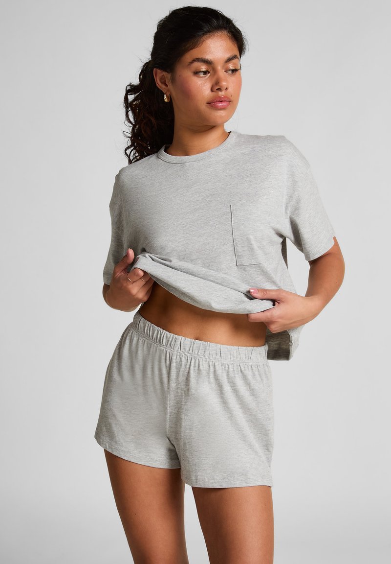 Ung kvinna som lyfter upp en grå kortärmad tröja för att visa magen, iklädd matchande grå shorts med elastisk midja framför en neutral bakgrund.