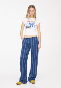 Hvid cropped t-shirt med blå grafik, parret med blå vertikalt stribede lette bukser. Skoene er gule med en brun sål.