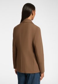 Blazer marrone su misura realizzato in tessuto strutturato, con colletto a rever e dettagli delle cuciture in evidenza, abbinato a pantaloni in denim blu.