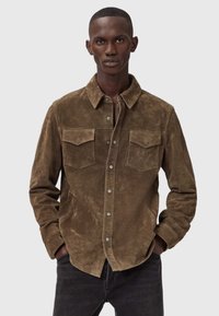 Man met kort haar in een bruine suède knoopshirt met borstzakken, handen in de zakken, staand tegen een effen lichte achtergrond.