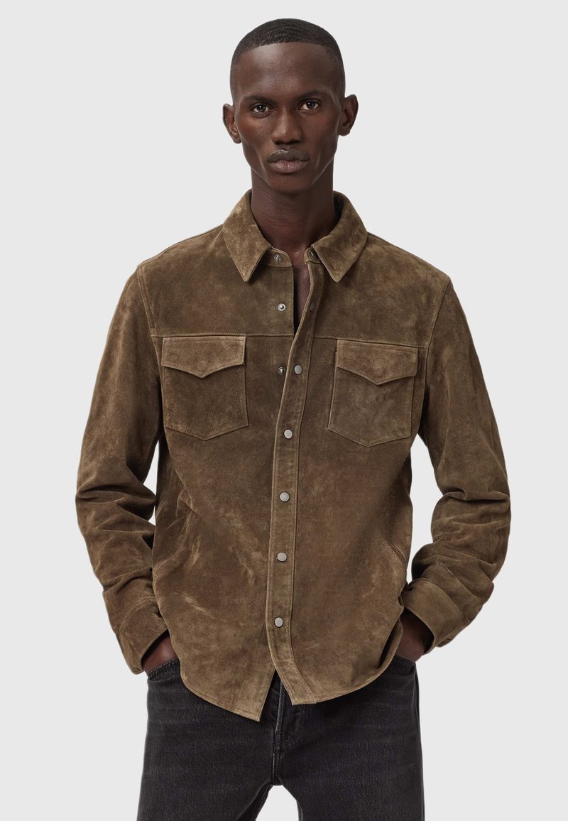 Man met kort haar in een bruine suède knoopshirt met borstzakken, handen in de zakken, staand tegen een effen lichte achtergrond.
