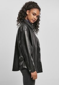 Veste en cuir noir avec un devant à boutons, un large col et une coupe ample. Elle possède des manches longues et des accents à boutons-pression.