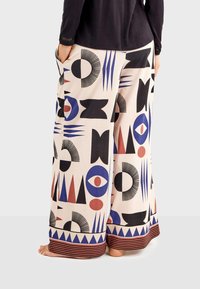 Wide-leg pants in light pink fabric featuring bold geometric patterns in black, red, and blue. Includes side pockets and striped hem.