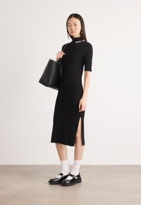 Abito aderente nero con maniche corte, collo alto e spacco laterale. Il modello tiene una borsa a tracolla nera, indossando scarpe nere e calzini bianchi.