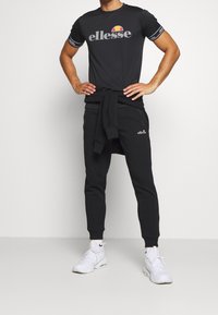 Černé sportovní tričko s šedým logem a barevným akcentem, v kombinaci s černými tepláky a bílými teniskami. Pohodlná látka, volný střih.