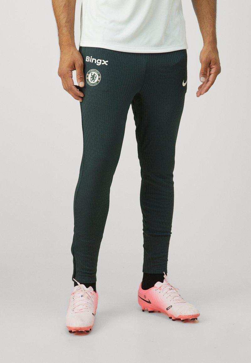 Pantalons de sport verts avec une texture côtelée, logo Nike et écusson de Chelsea FC. Ils présentent une coupe slim et des bandes contrastantes sur les côtés.