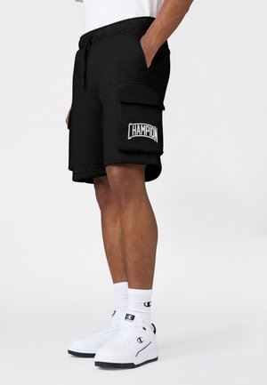 Man draagt zwarte Champion shorts met zijzak en witte Champion sneakers en kousen tegen een effen achtergrond.