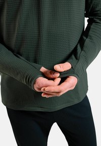 Langärmliges, dunkelgrünes Hemd aus strukturiertem Stoff mit Daumenlöchern an den Bündchen. Kombiniert mit schwarzen Hosen. Hände sind ineinander gelegt.