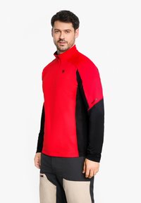 Rot-schwarzes Pullover-Oberteil mit halbem Reißverschluss und langen Ärmeln, das eine glatte Textur und ein Logo-Detail auf der Brust und dem Ärmel aufweist.